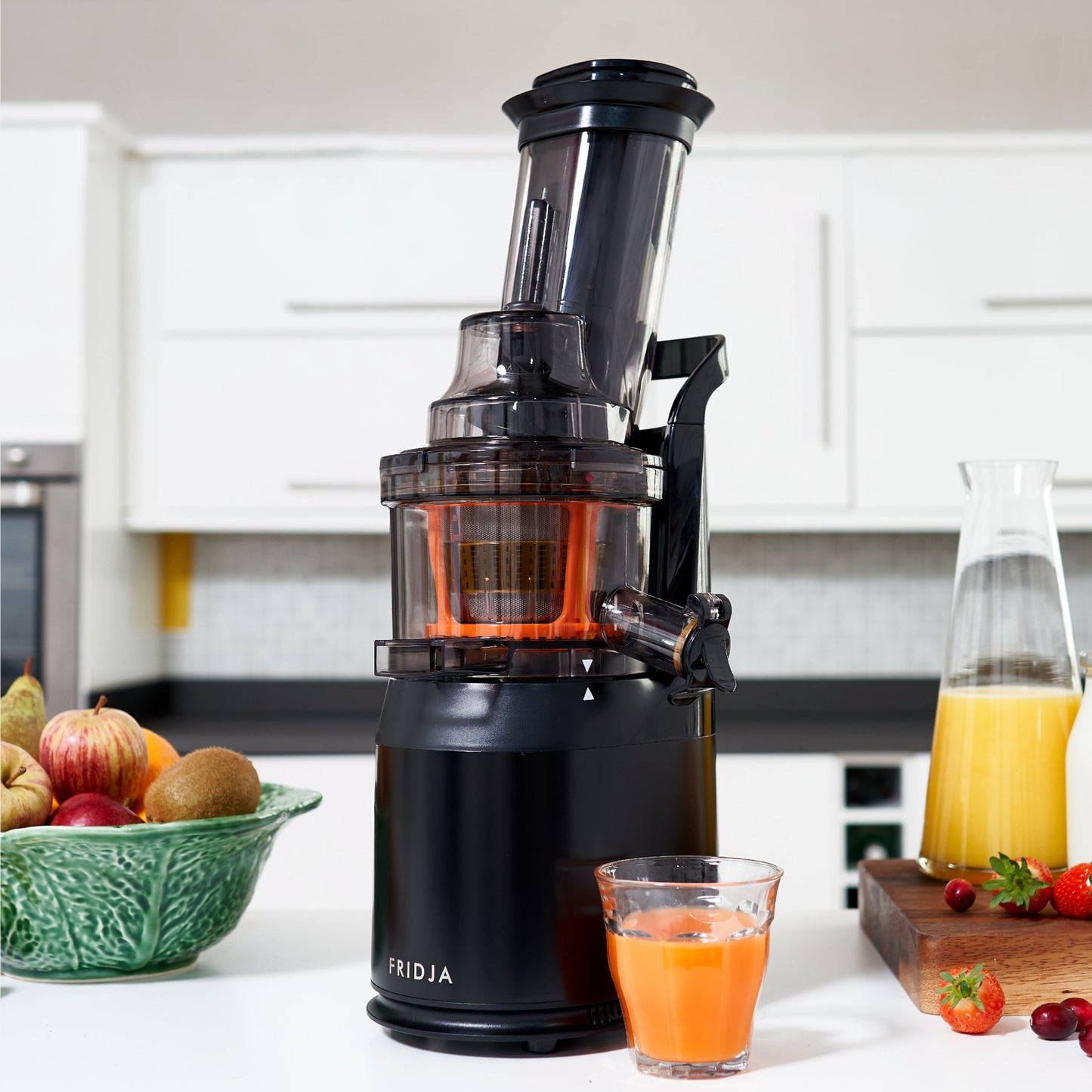 A black Fridja f1900 Whole Fruit Juicer stands on a kitchen counter, filling a glass with juice. Nearby are assorted fruit in a bowl, a carafe of juice, and strawberries on a cutting board in front of white cabinets.
