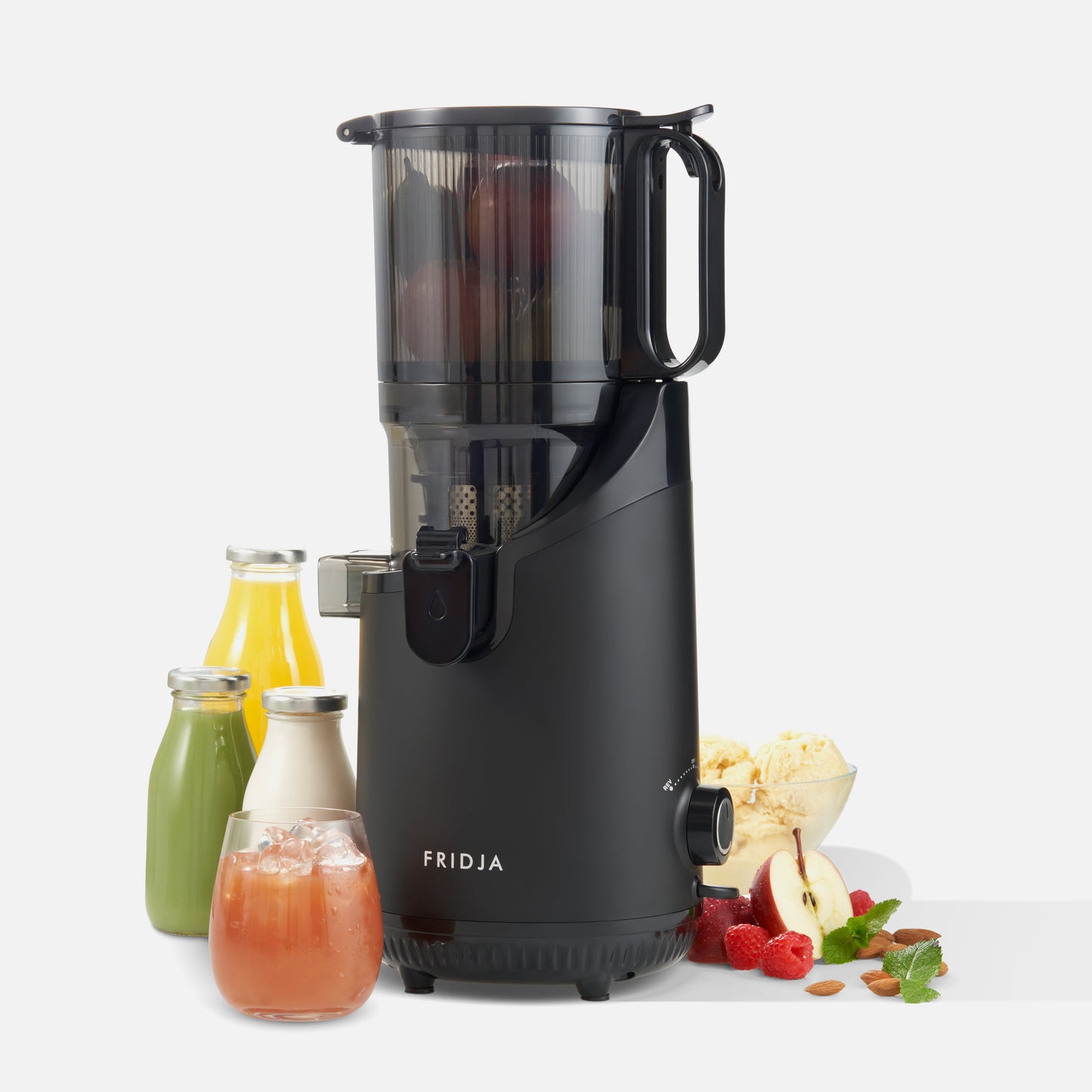 A black Fridja f2500 Self Feeding Whole Fruit Juicer, easy to clean, sits on a white background with bottles and a glass of cold-pressed juice, fresh fruits, herbs, ice cream, and apples visible inside.