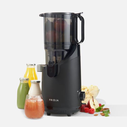 A black Fridja f2500 Self Feeding Whole Fruit Juicer, easy to clean, sits on a white background with bottles and a glass of cold-pressed juice, fresh fruits, herbs, ice cream, and apples visible inside.