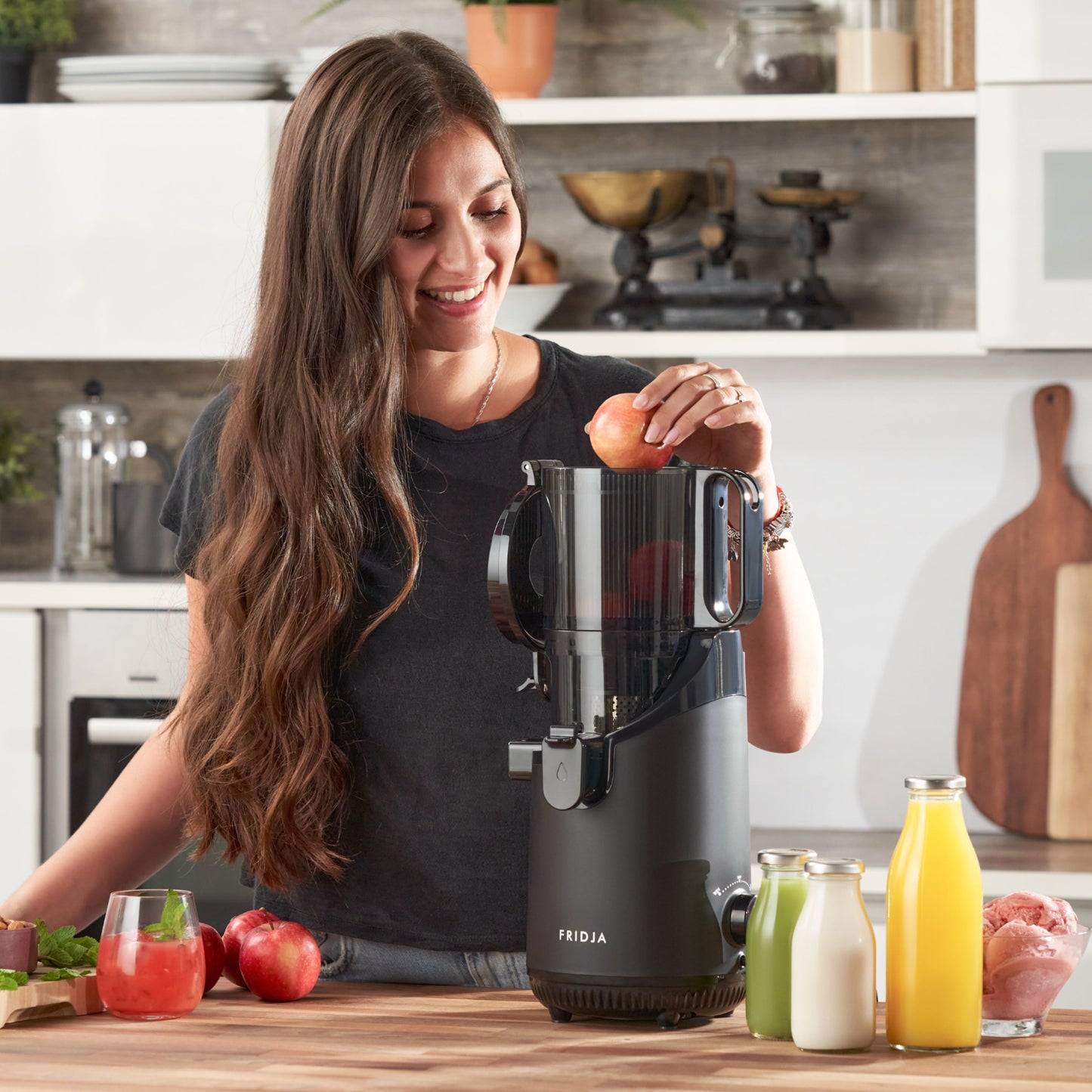 A woman smiles in a bright kitchen as she prepares to place an apple into a black Fridja f2500 Self Feeding Whole Fruit Juicer. Bottles of cold-pressed juice and fresh fruits and vegetables sit on the counter nearby.
