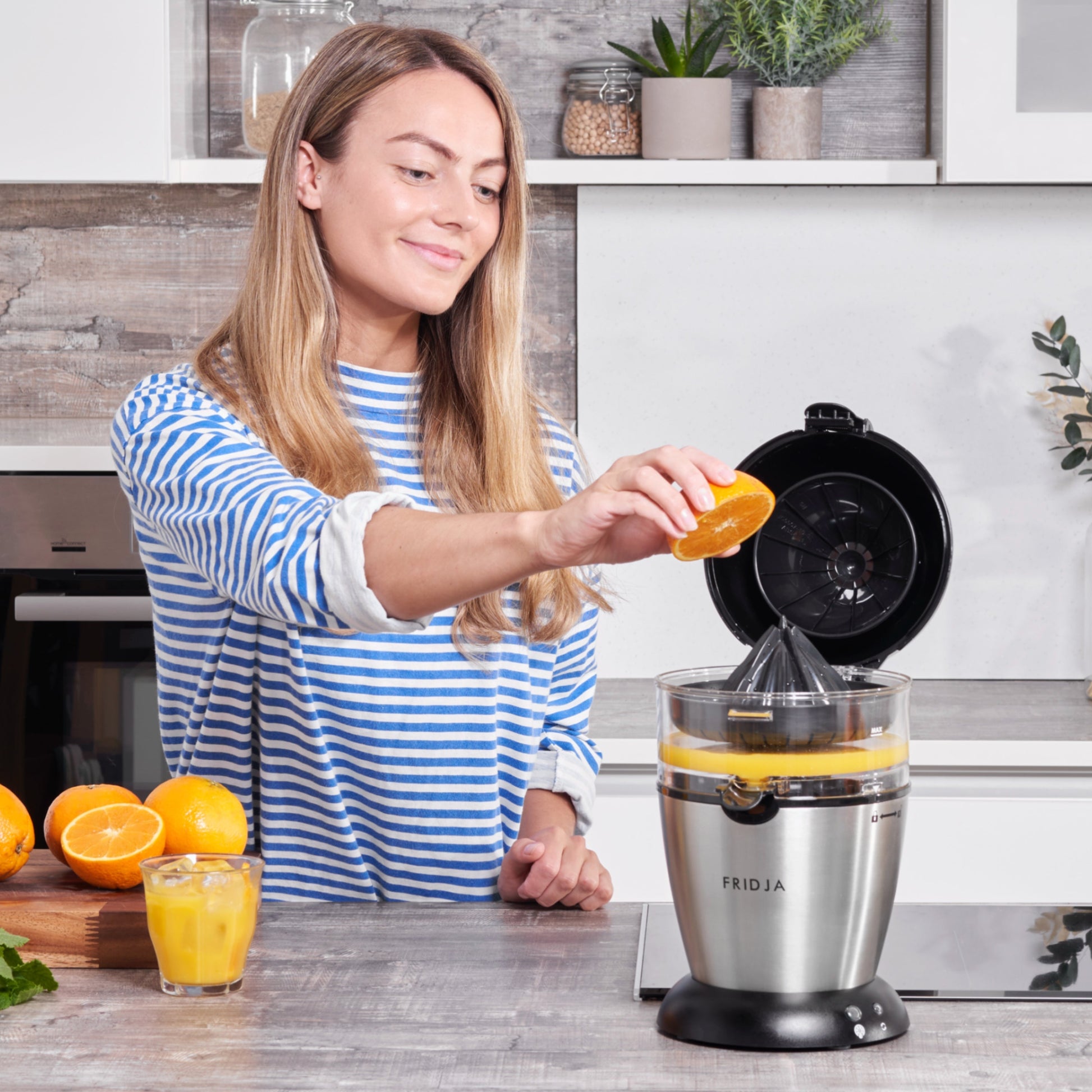 A woman in a blue and white striped shirt uses the Fridja f900 Automatic Citrus Juicer - Refurbished Grade A to squeeze an orange in a modern kitchen, with a glass of juice and more oranges on the counter beside her.